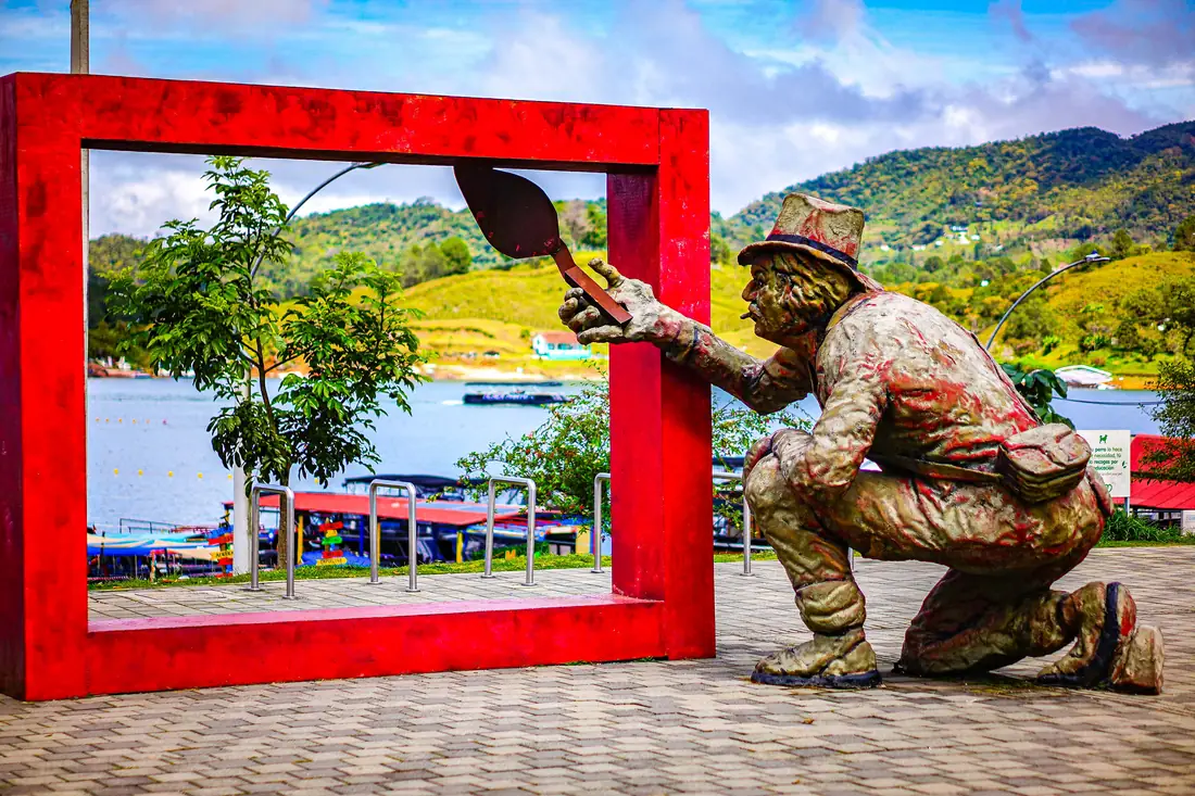 Sculpture of a zocalero painting, framed in red, with the reservoir in the background.